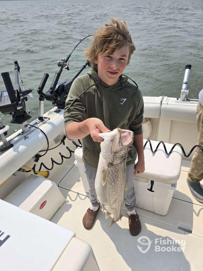 Young angler proudly holding a large Pike while fishing on a boat, showcasing a successful catch in a scenic waterway.