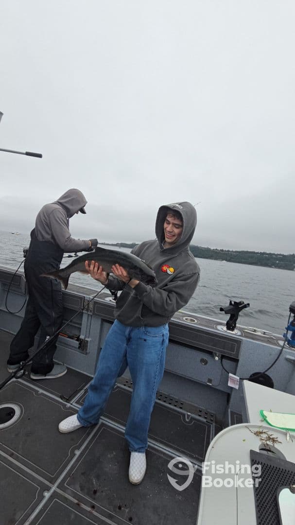 A young angler joyfully holding a sizable fish aboard a fishing boat, showcasing the excitement of a successful catch on a cloudy day.