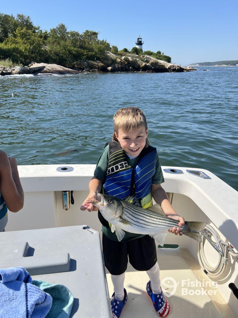 A young angler proudly displaying a striped bass while fishing on a boat, with a scenic lighthouse in the background.