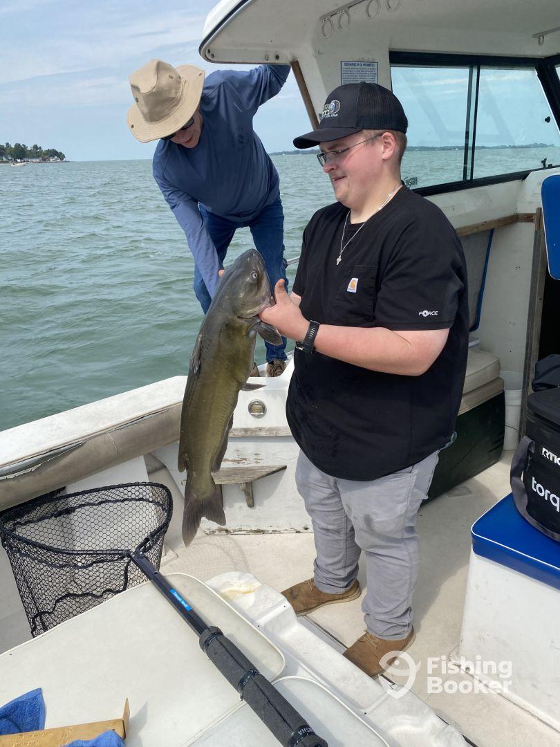 A young angler proudly displaying a large catfish while fishing on a boat, with an adult assisting in the background.