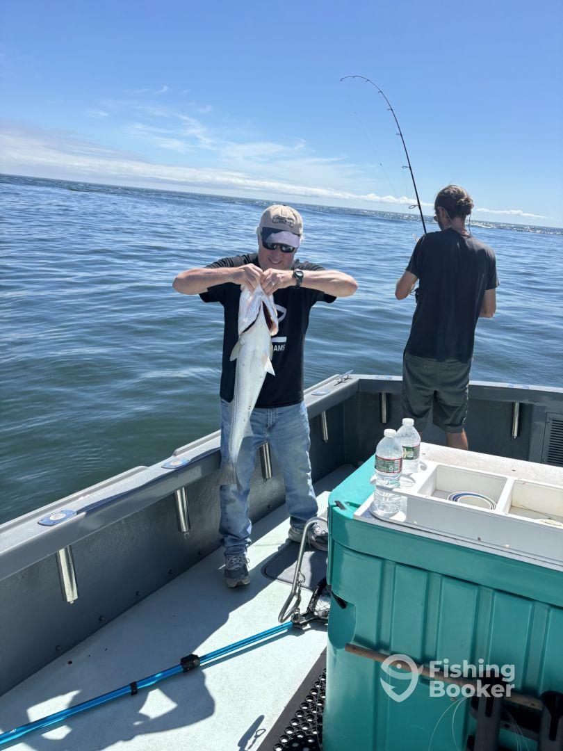 An angler proudly displaying a large fish on a boat during a fishing trip, with another person actively fishing in the background.