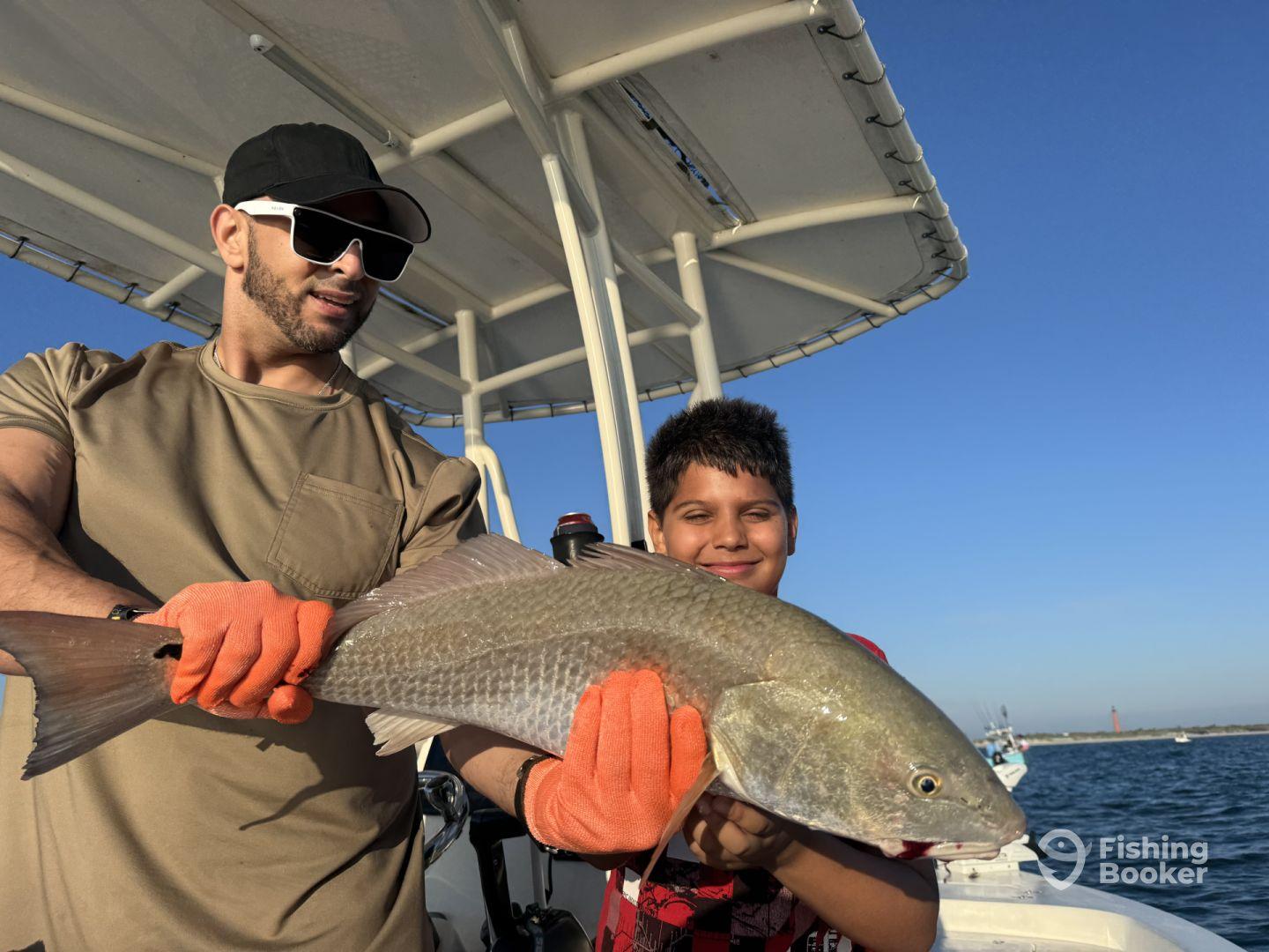 A young angler and an adult proudly displaying a large Redfish on a sunny day out on the water, showcasing a successful fishing trip.