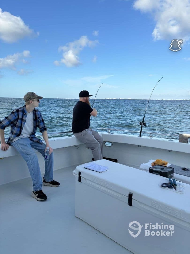 Two anglers engaged in fishing on a charter boat, with one actively reeling in a line while the other observes, set against a scenic ocean backdrop.