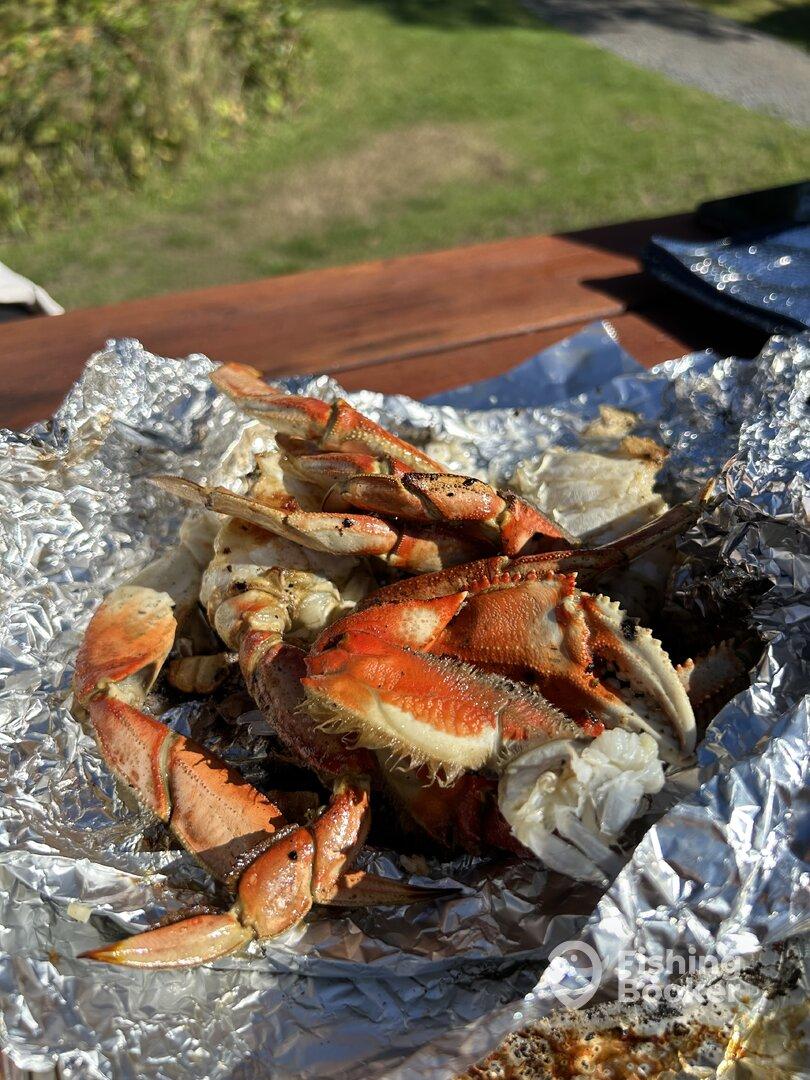 A close-up view of grilled crab claws on aluminum foil, set on a wooden table in a natural outdoor setting.