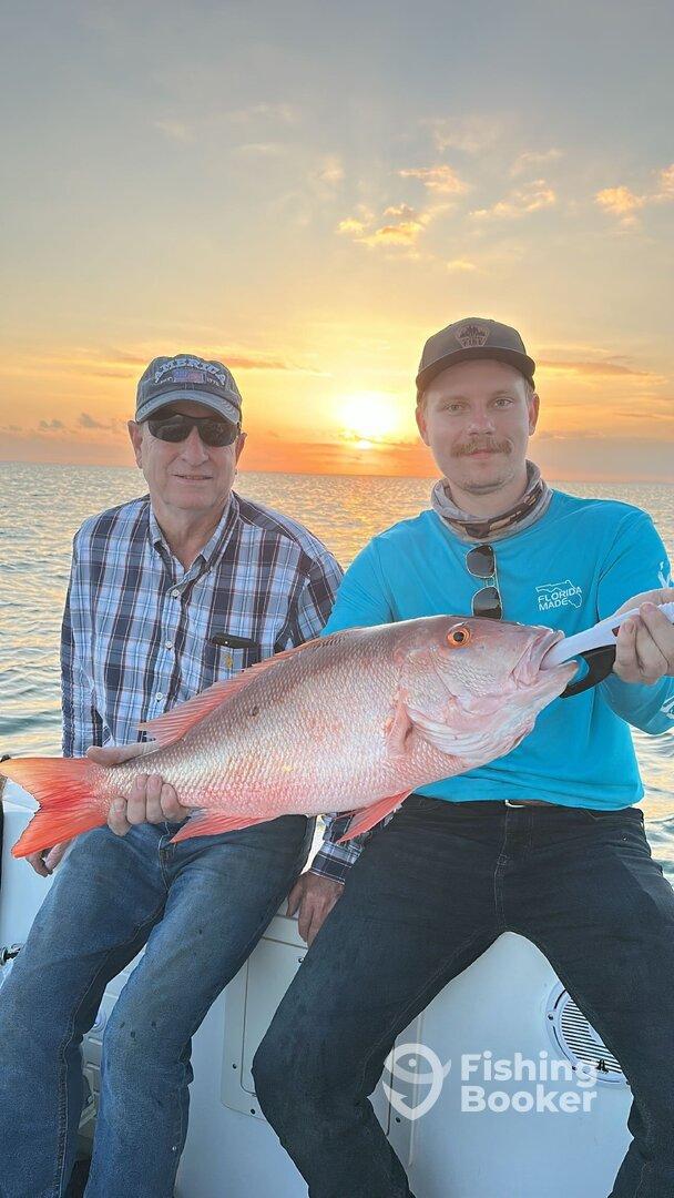 Two anglers proudly displaying a large Snapper against a stunning sunset backdrop while out on the water.
