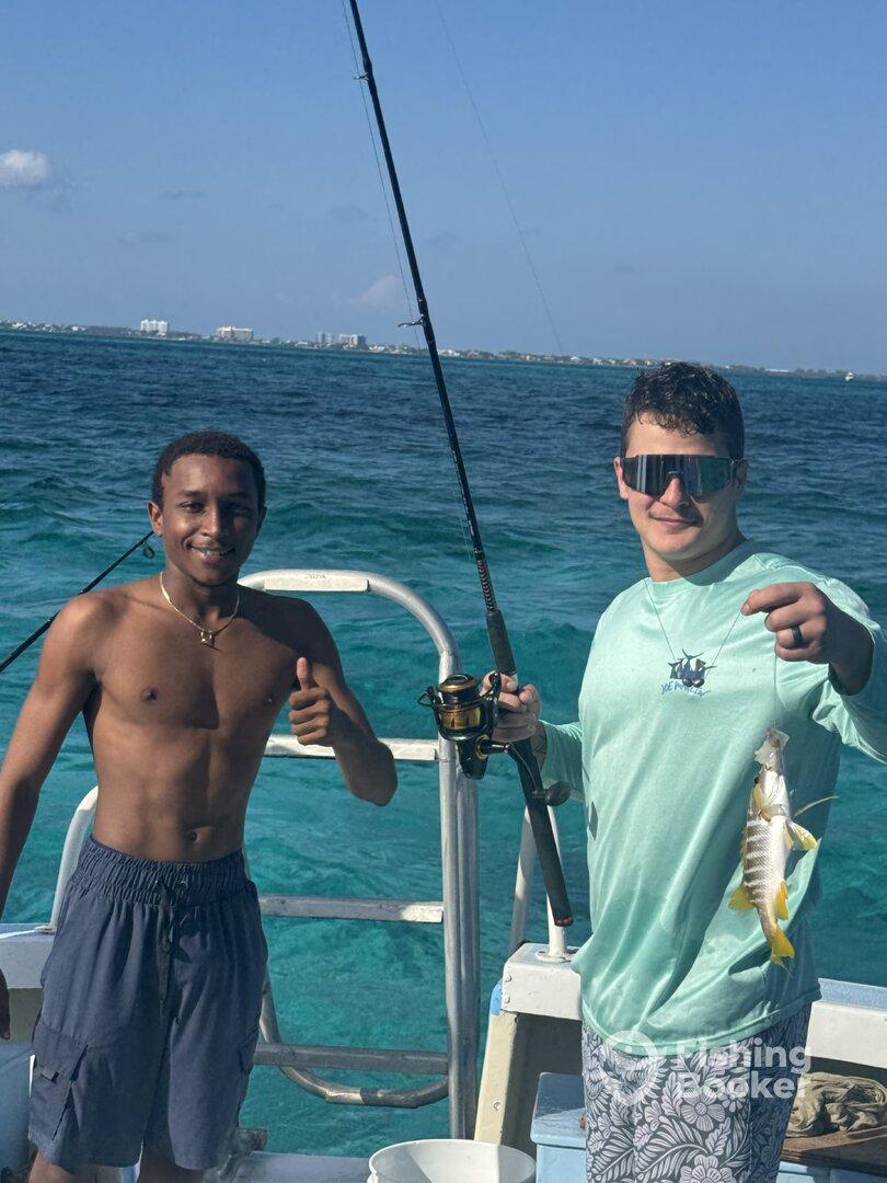 Two young anglers on a boat, one proudly displaying a small fish while the other gives a thumbs up, showcasing a fun day of fishing in clear blue waters.
