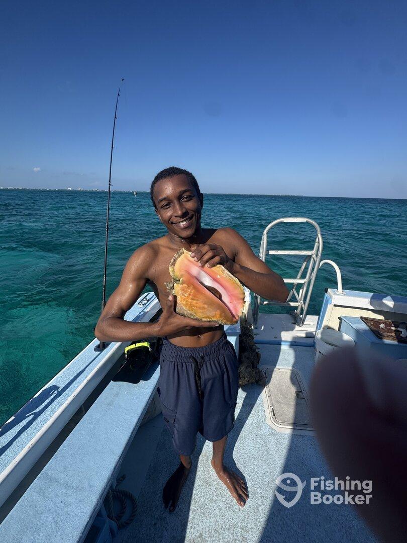 A young angler proudly displaying a large conch shell while on a fishing trip in clear turquoise waters.