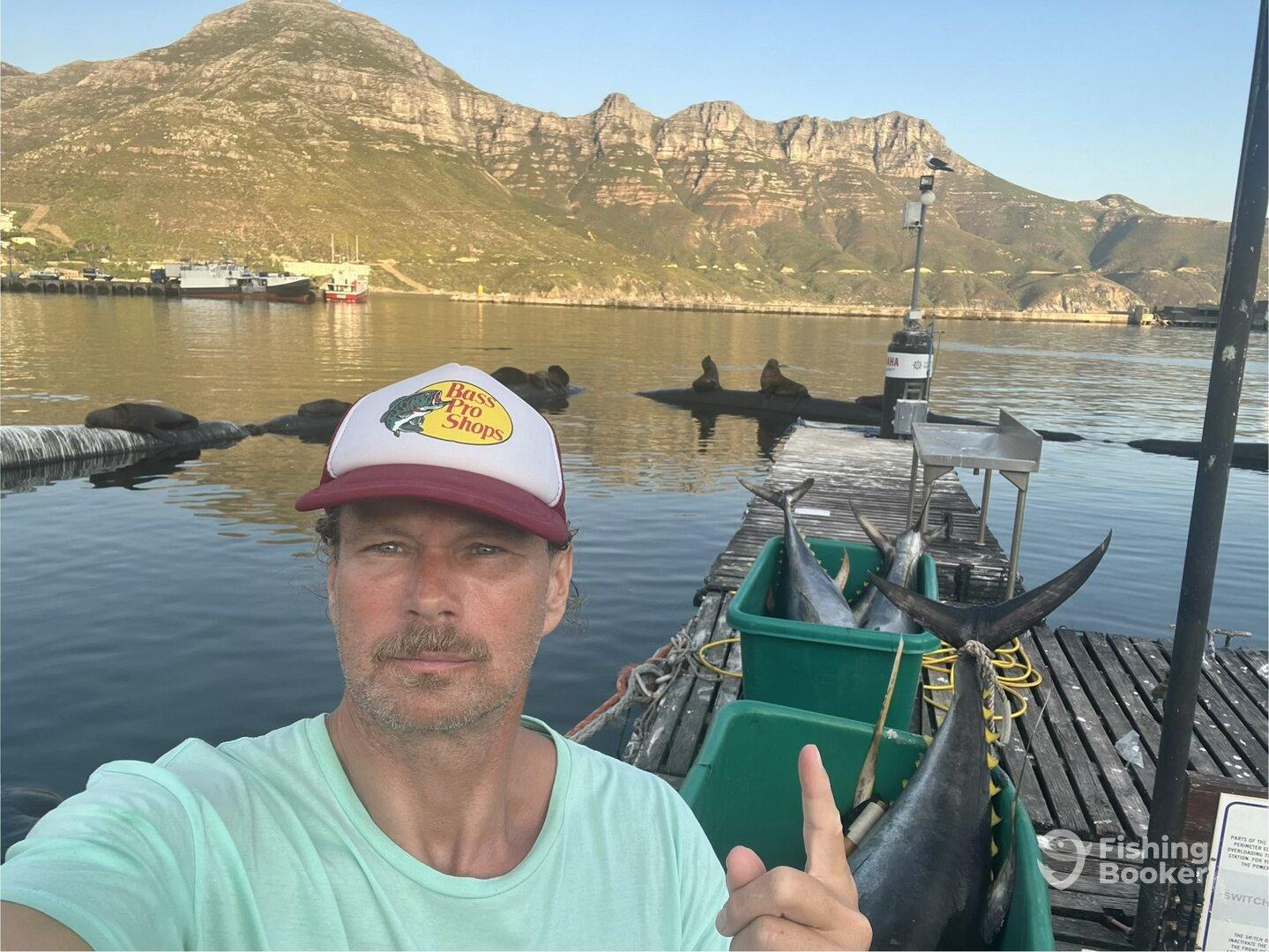A fisherman poses near large fish stored in green bins at a dock, with scenic mountains in the background.