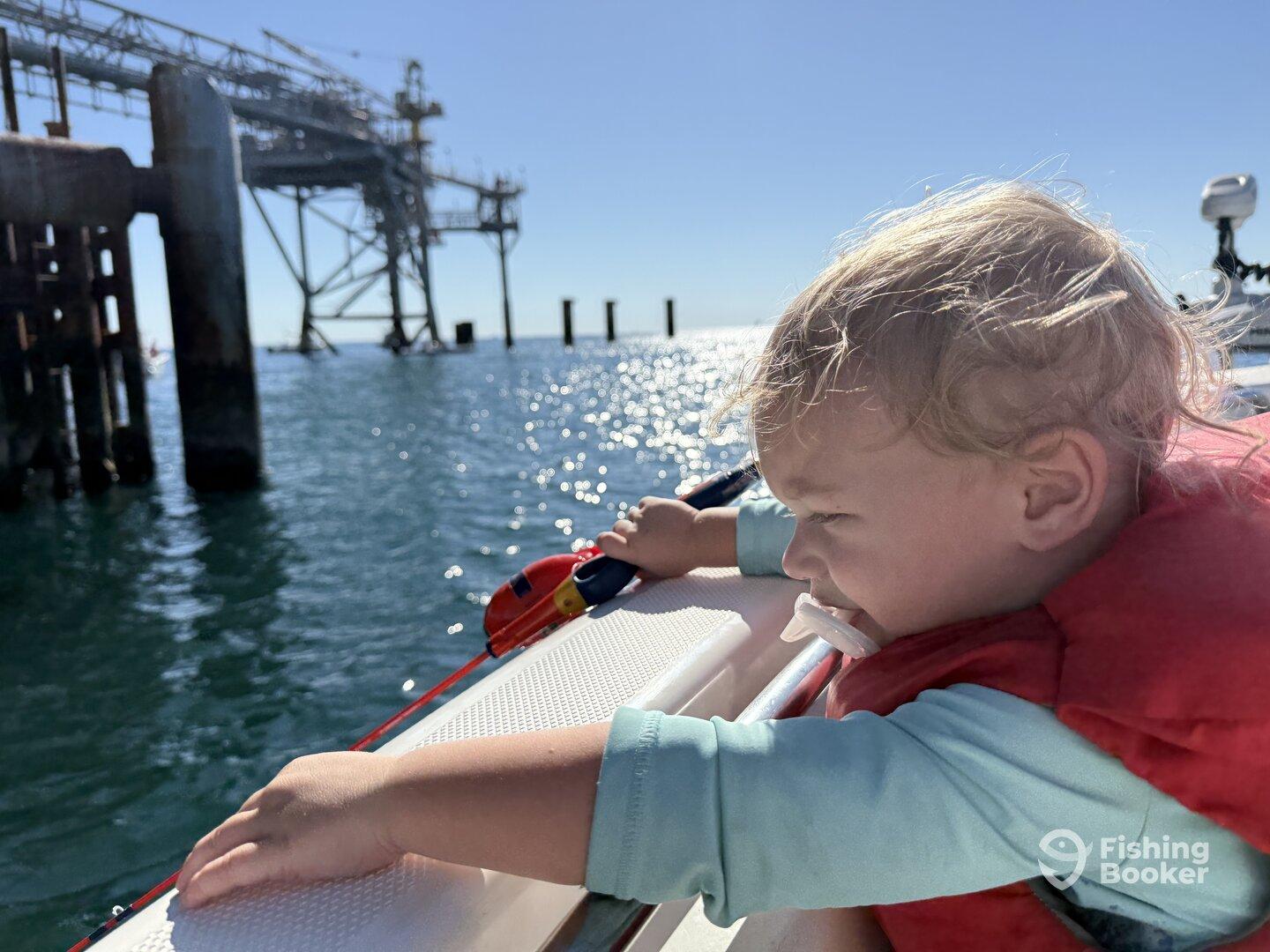 A young child in a life jacket gazes out from a boat towards a fishing pier, enjoying a sunny day on the water.
