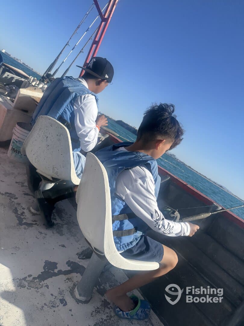 Two young anglers seated on a boat, focused on fishing in a sunny coastal environment.