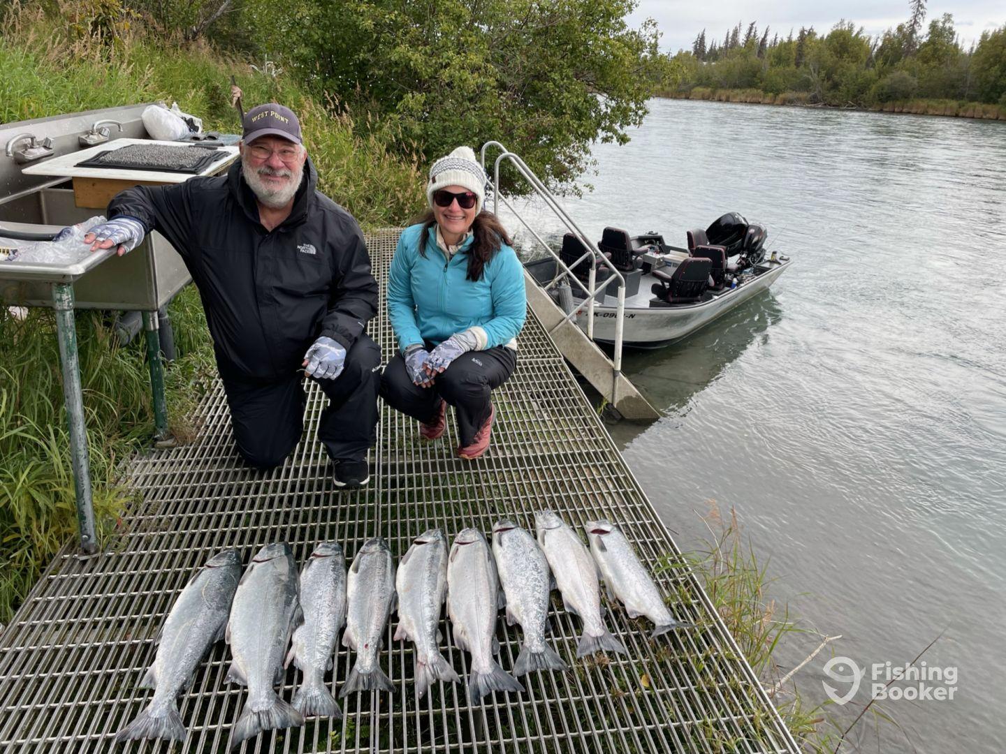 Two anglers proudly displaying their catch of salmon on a dock by a river, with a fishing boat visible in the background.
