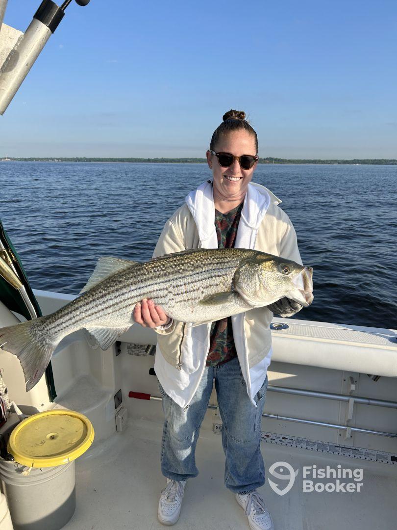 Angler proudly displaying a large Striped Bass while fishing on a boat, showcasing a successful day on the water.