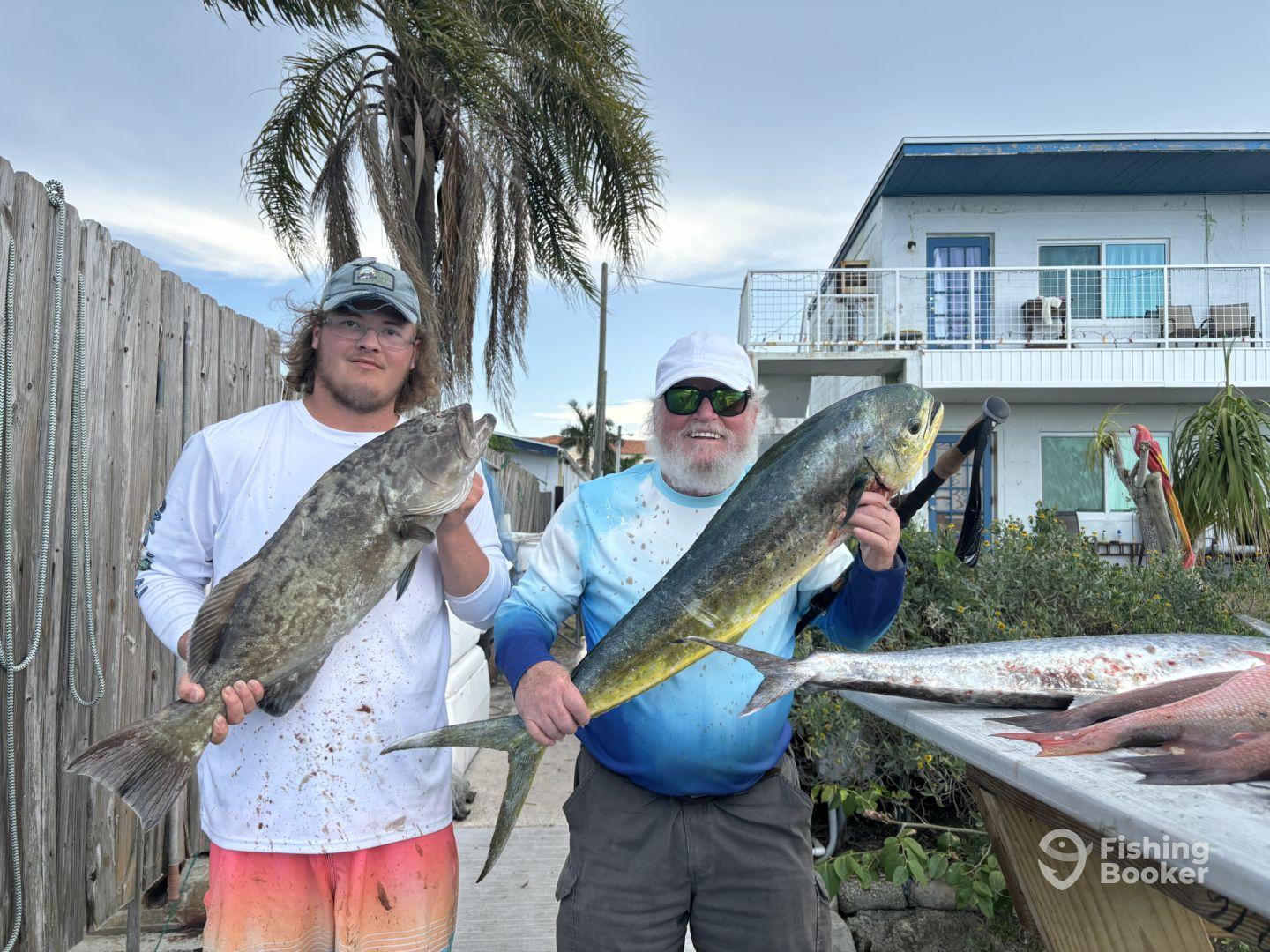 Two anglers proudly displaying their catches, a large Grouper and a Mahi Mahi, at a residential dock after a successful fishing trip.