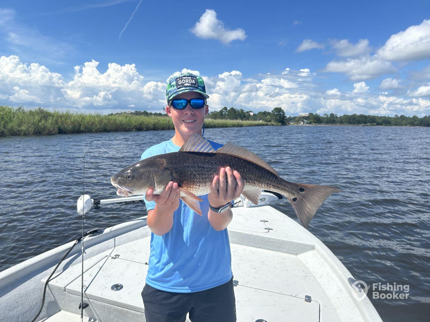 Young angler proudly holding a large Redfish while fishing on a boat in a scenic waterway.