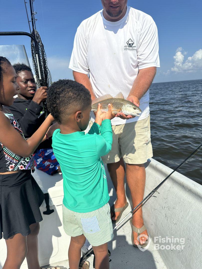 A young boy excitedly holds a fish with the help of an adult, surrounded by family on a fishing trip.