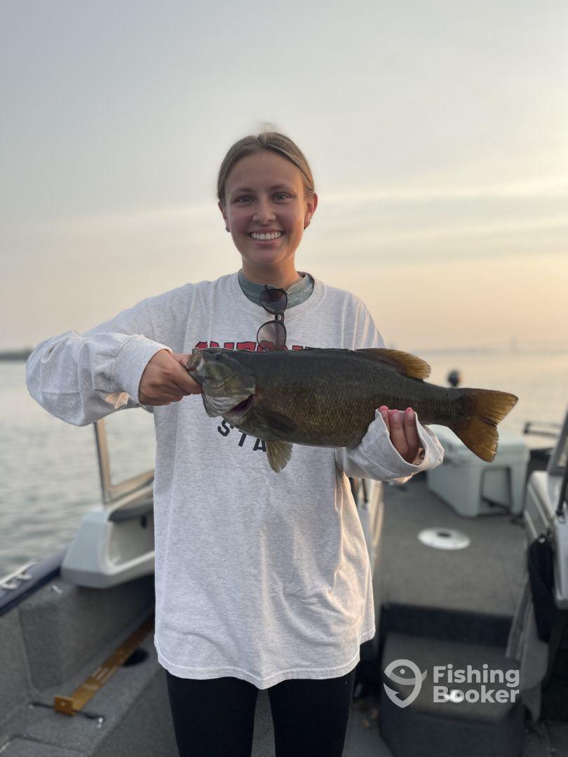 A young woman proudly displaying a large Smallmouth Bass while fishing on a boat during sunset.