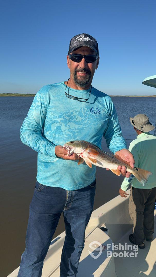 Angler proudly displaying a Redfish while fishing on a calm river, with another fisherman in the background.