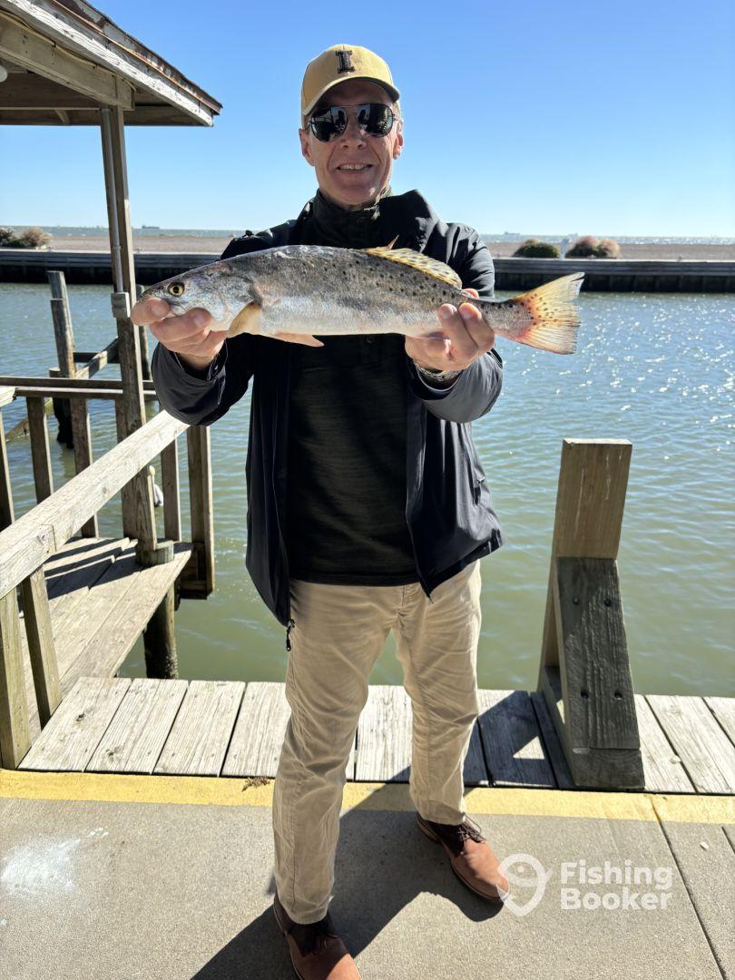 An angler proudly displaying a large Speckled Trout while standing on a dock, showcasing a successful fishing trip.