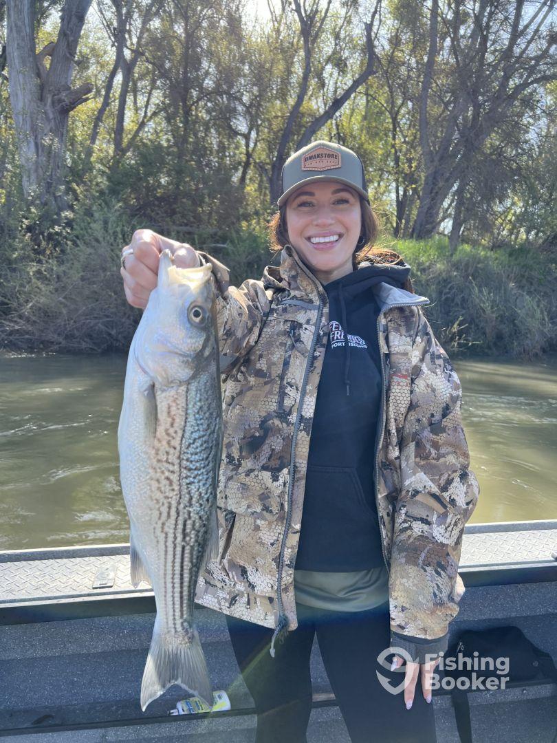 A smiling angler proudly displays a large striped bass while fishing on a calm river, showcasing a successful day on the water.