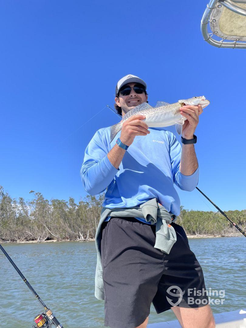 An angler proudly displaying a Speckled Trout while fishing on a sunny day, surrounded by lush greenery along the waterway.