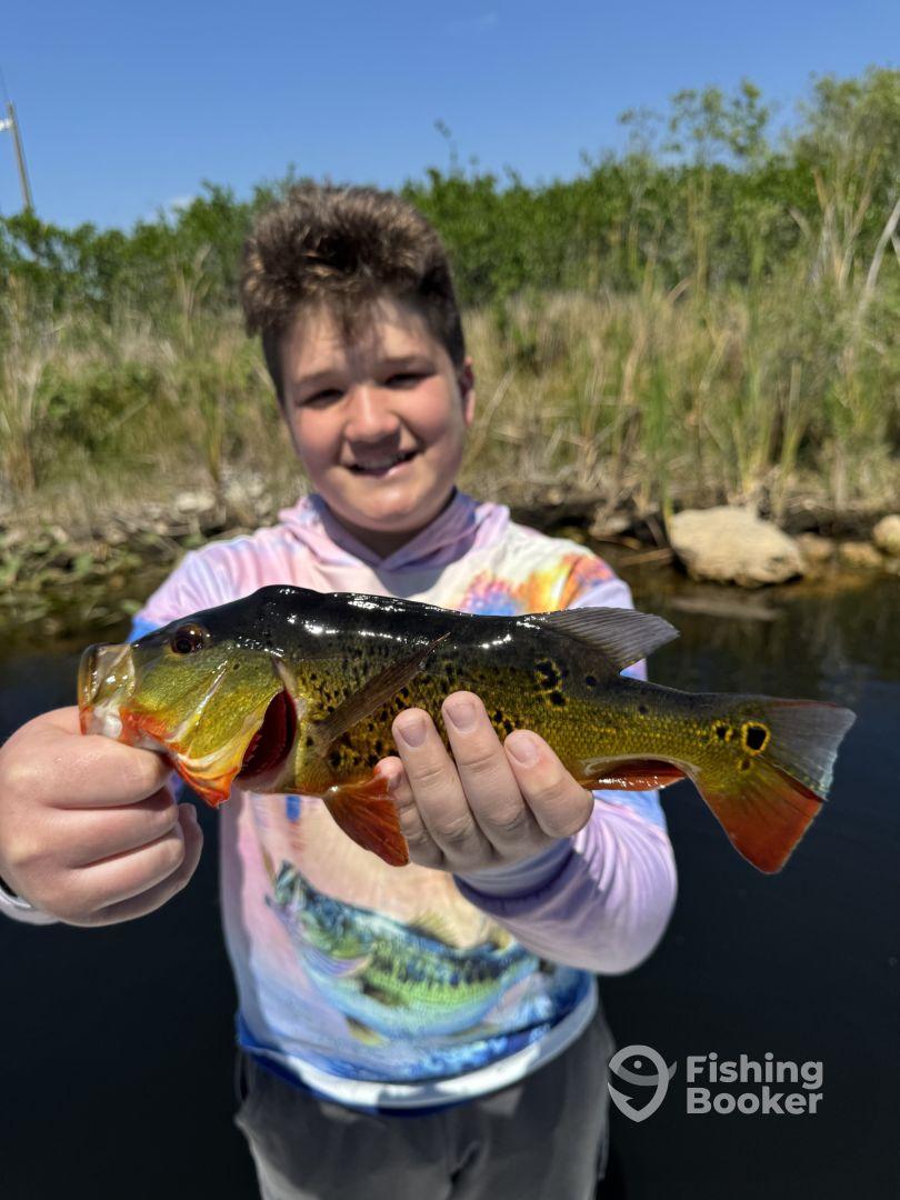 A young angler proudly displaying a vibrant Peacock Bass caught during a fishing trip in a scenic waterway.