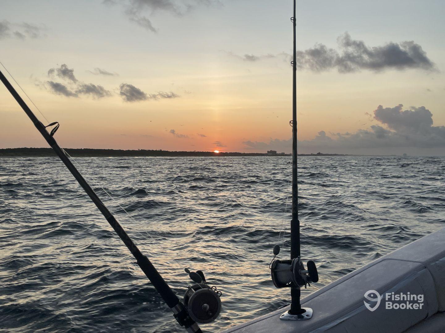 A serene sunset view from the side of a fishing boat, showcasing fishing rods against the backdrop of a tranquil ocean.