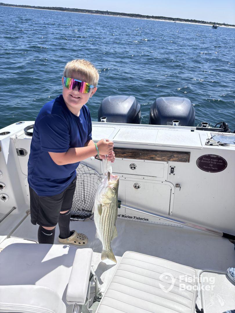 A young angler proudly displaying a large striped bass aboard a boat, enjoying a sunny day on the water.