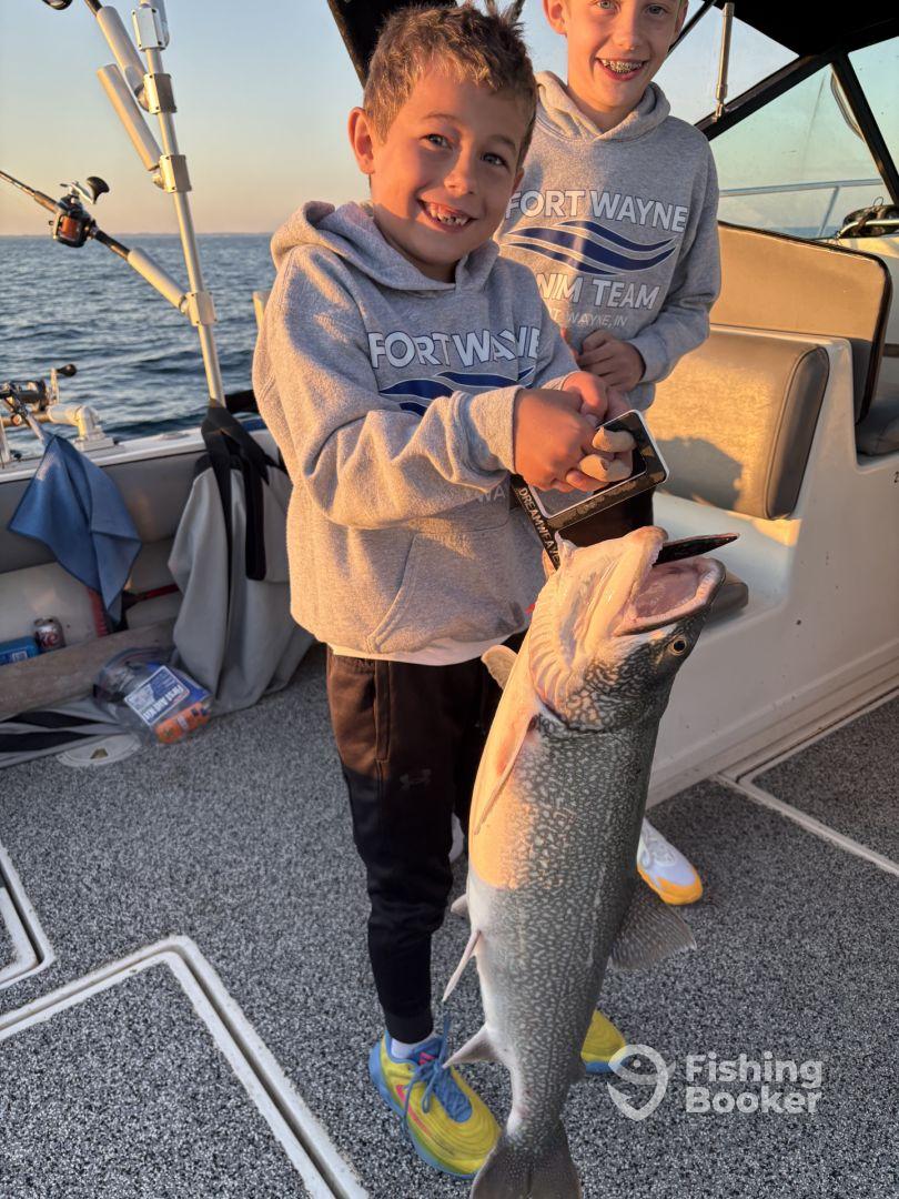 Two young anglers proudly displaying a large Lake Trout aboard a fishing boat during a sunset outing.
