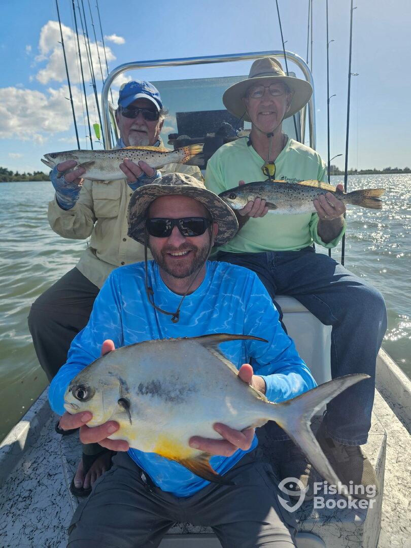 Three anglers proudly displaying their catch, including a large Pompano and Speckled Trout, while enjoying a sunny day on the water.