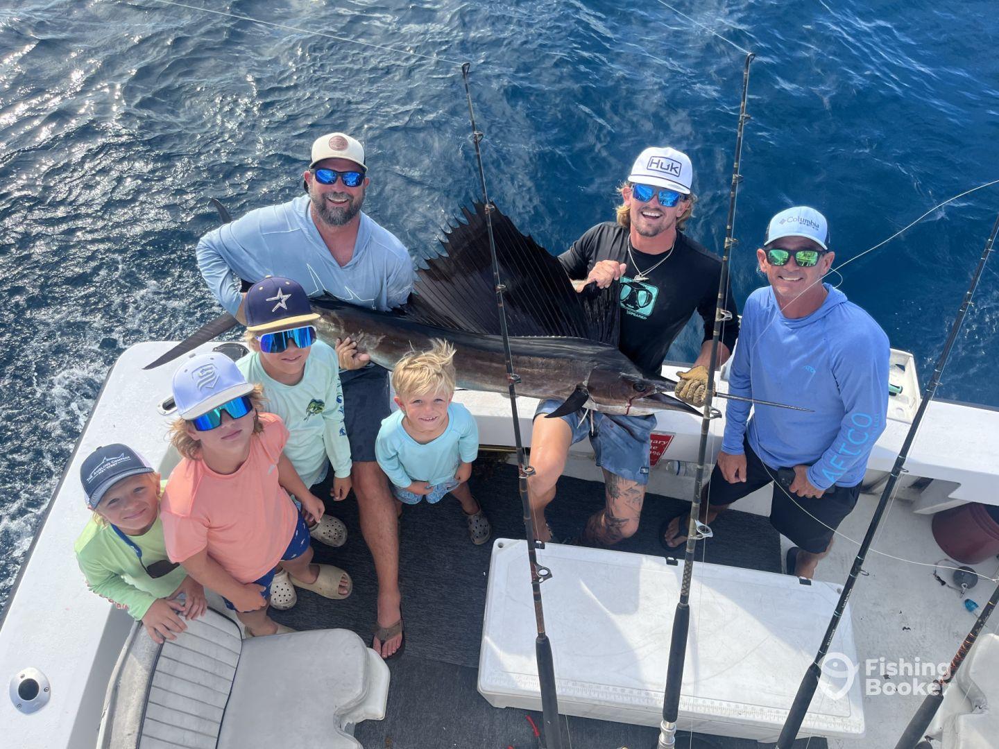 A group of anglers, including children, proudly displaying a large Sailfish on a fishing boat, showcasing a successful deep-sea fishing trip.