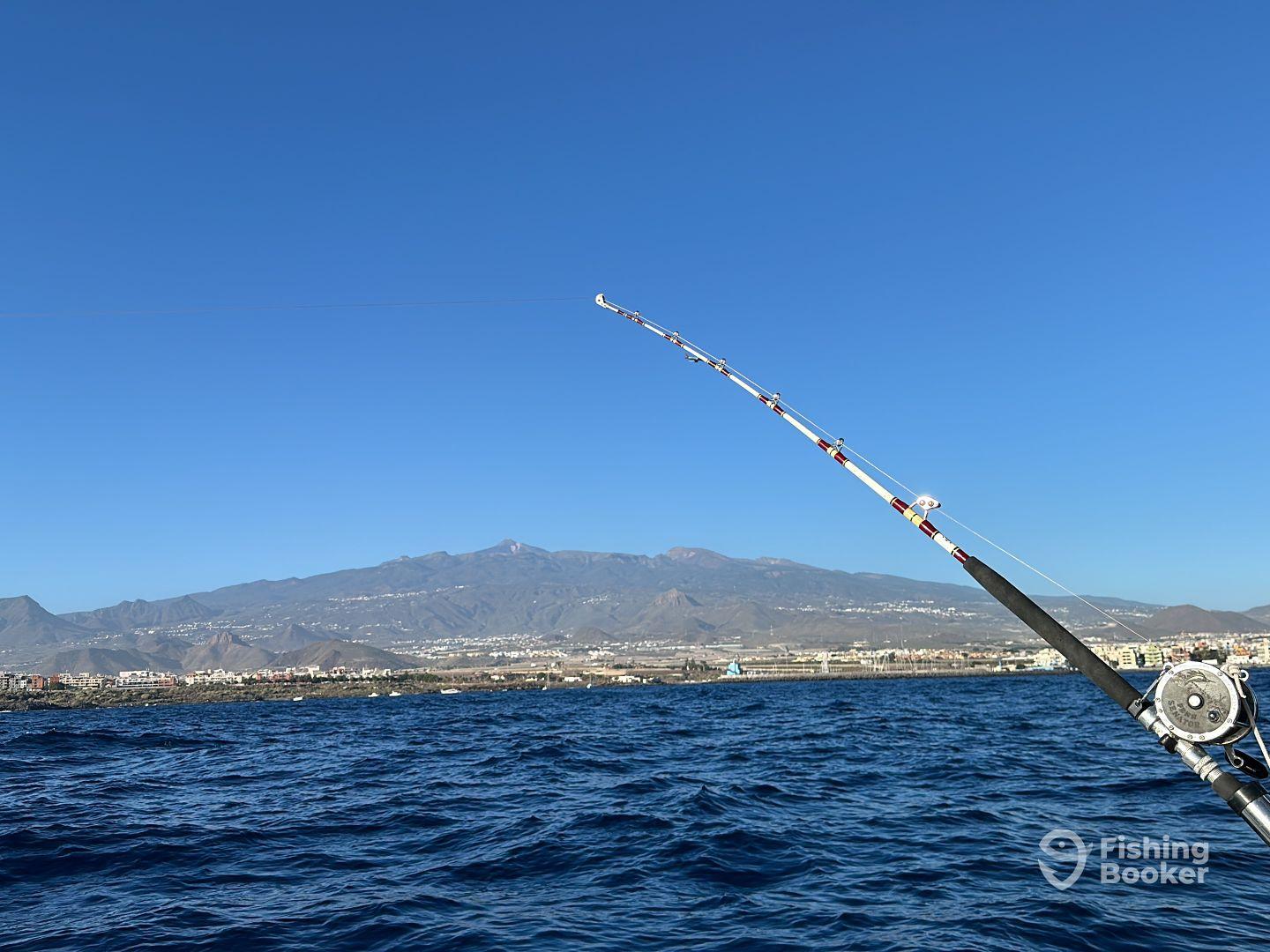 Fishing rod in action against a scenic backdrop of mountains and coastline, showcasing a vibrant blue sky and ocean.