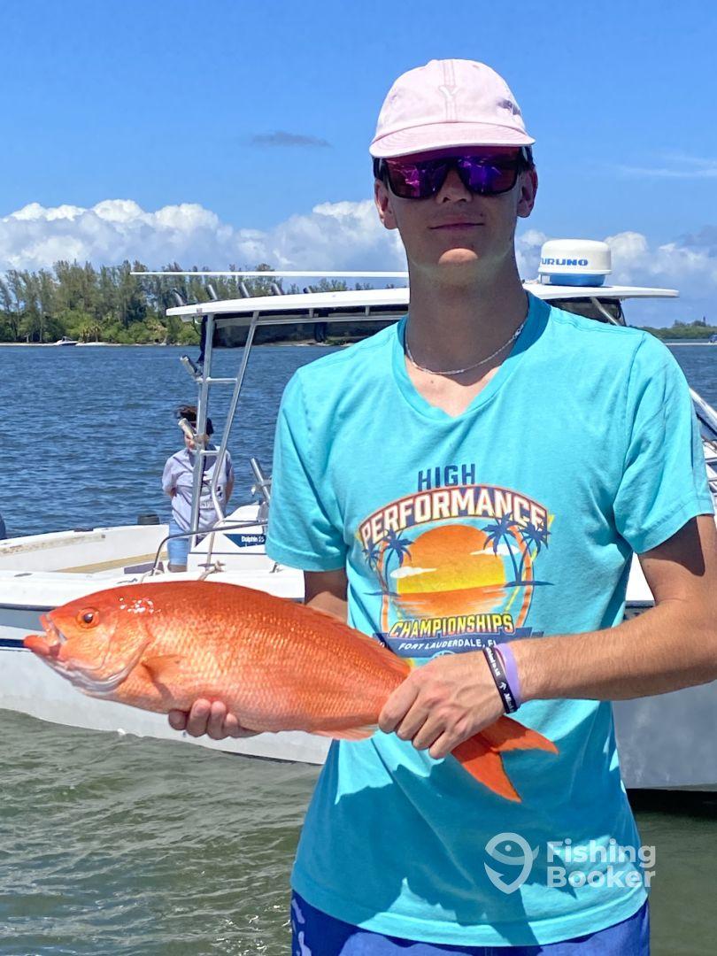 A young angler proudly displaying a vibrant Red Snapper while standing near a boat in a sunny coastal setting.