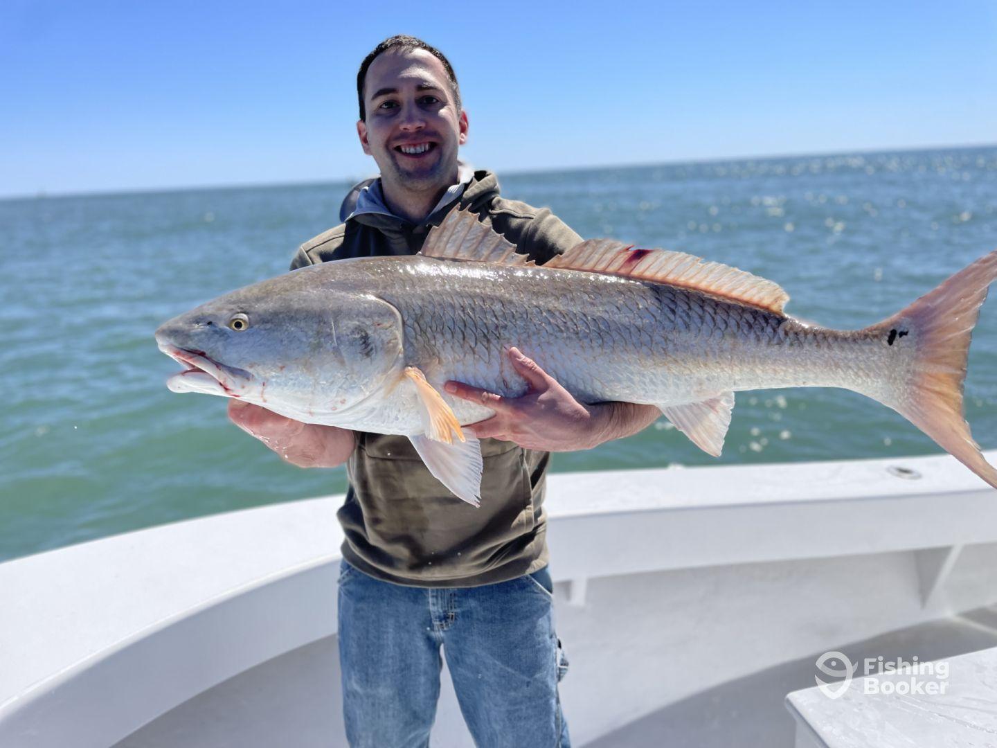An angler proudly displaying a large Redfish while aboard a fishing charter, with a clear blue sky and water in the background.
